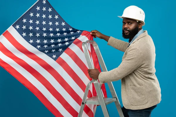 Young bearded African-american worker in protective helmet and formalwear standing on stepladder and holding US flag