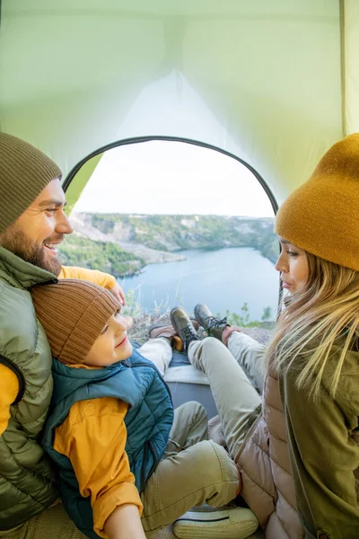 秋の週末の旅行中に湖の前でテントの中で休んでいる家族がいる間 若い女性は父によって座っている小さな息子に話します — ストック写真