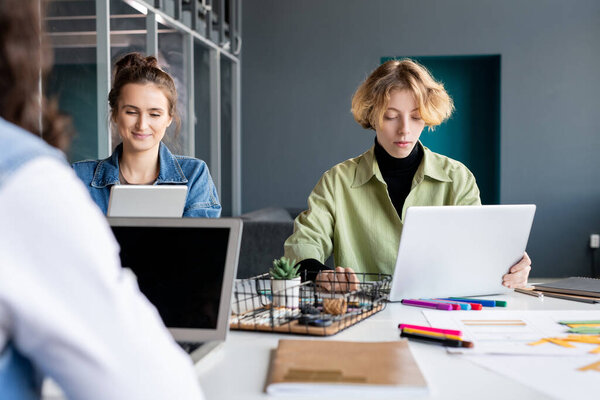 Two young creative female programmers working over new software in front of laptops while sitting by desk in front of colleague