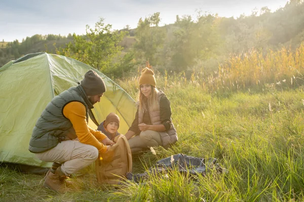 年轻人蹲在帐篷里 一边从背包里拿出食物 一边与妻子和儿子一起在周末的旅行中休息和吃午饭 — 图库照片