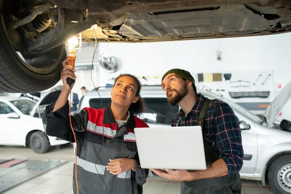 Young female mechanic with lamp and her male colleague with laptop looking at bottom of broken car to find out the reason of problem