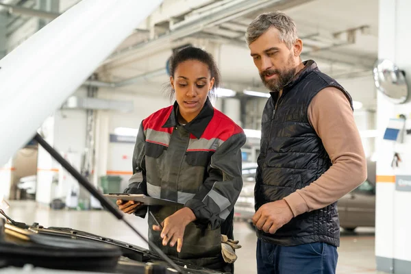 自動車修理サービスの若い女性技術者は ワークショップで男性クライアントをコンサルティングしながら オープンエンジンコンパートメントの部品の一つを指しています — ストック写真