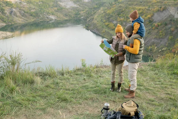 年轻的三口之家站在草地上 面对自然环境中的河流或湖泊 一边看着地图 一边想着下一步该去哪里 — 图库照片
