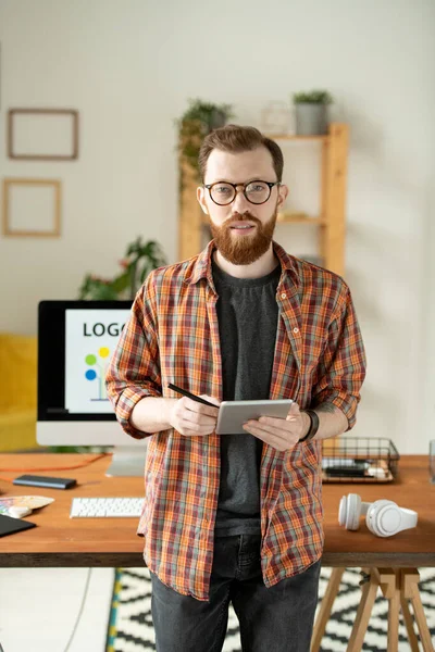 Young bearded creative designer standing by table in front of camera and looking at you while using tablet and stylus against computer monitor