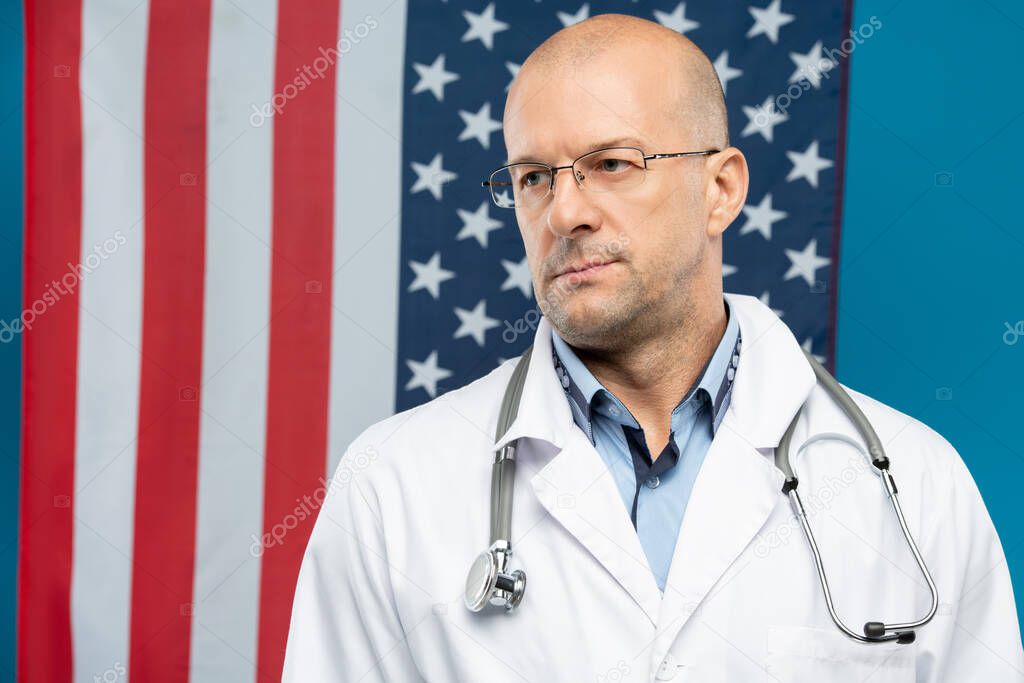 Contemporary bald clinician in eyeglasses and whitecoat standing in front of camera against stars-and-stripes and blue background