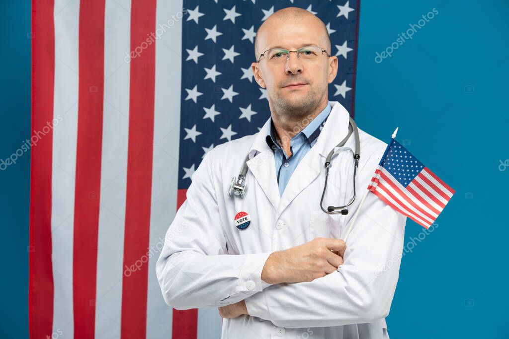 Confident middle aged doctor in eyeglasses and whitecoat holding small American flag while standing against stars-and-stripes background