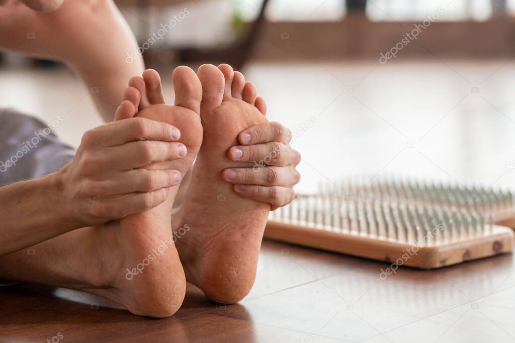 Barefoot man sitting on wooden floor with yoga therapy pads with metallic nails near by and holding his feet in hands during one of exercises