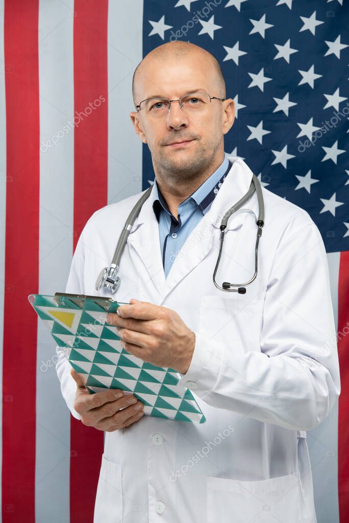 Middle aged contemporary doctor in eyeglasses holding clipboard with document while standing against stars-and-stripes in front of camera