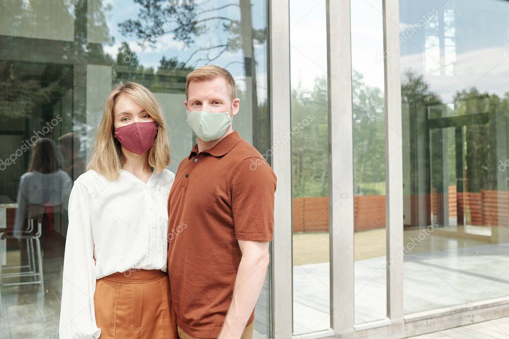 Beautiful couple in cloth masks standing against cottage and embracing while posing for photo