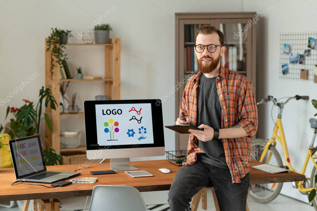 Cheerful male freelance designer holding stylus over tablet while sitting on table, drawing sketches of new logo and looking at you with smile