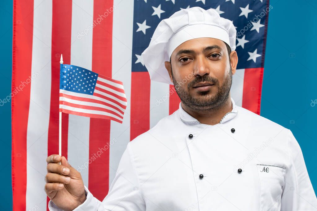 Young serious mixed-race chef in white uniform holding us flag while standing in front of camera against stars-and-stripes background