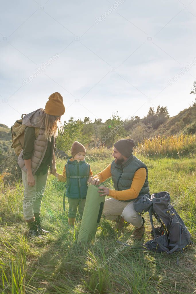 Young couple and their cute little son with backpacks standing on green grass in front of camera while man unrolling rug to have some rest