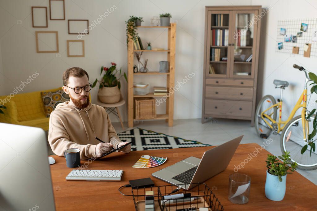 Young bearded freelance designer looking at laptop display while using tablet, stylus and color palette for drawing logo or something else