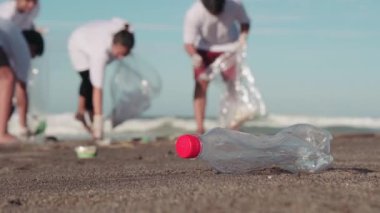 Seçici odak noktasında plastik şişe düşük kesimli bıçak atışı. Bir grup genç gönüllü, kirli plajdan deniz kenarındaki özel çöp torbalarına kadar diğer çöpleri topluyor.