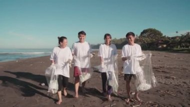 Kirli sahil boyunca çevreyi korumaya yardım eden çöp torbalarıyla kameraya doğru yürüyen neşeli gönüllülerin tam görüntüsü.
