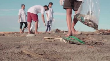 Düşük kesimlerde bir adam kumlu sandalyede yürürken ve plaj bölgesini temizlerken takımının da çöpleri topladığı özel bir çöp torbasına koyarken çekilmiş.