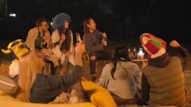 Neşeli bir grup arkadaş komik şapkalar takıp çevrimiçi çeviri izliyorlar tezahürat yapıyorlar, ilahi söyleyip bira içiyorlar açık havada iyi vakit geçiriyorlar.