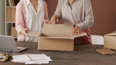 Çevrimiçi giyim mağazasında masanın yanında duran çok ırklı genç bayanları ve paketleme siparişlerini yukarı kaldır. Giysileri karton kutuya koyup mühürlüyorlar.