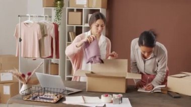 Çok ırklı genç bayanların online giyim mağazasının ofisinde masanın başında beklemeleri ve katlanmış giysileri karton kutuya koyup teslimata hazırlanmaları.