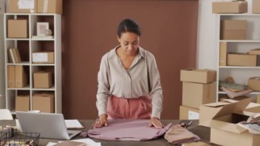 Küçük çevrimiçi kıyafet mağazasının ofisinde duran mutlu siyahi genç bir kadının portresini izlemek ve tişörtün üzerindeki kırışıklıkları düzeltmek, sonra da kameraya bakmak ve gülümsemek.