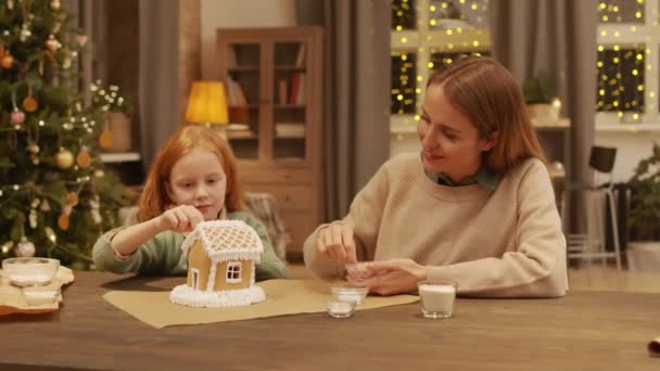 中ショットの幸せな小さな女の子飾る大きなクリーミーなジンジャーブレッドハウスとともに彼女のお母さん取得準備のためにクリスマスのお祝い — ストック動画