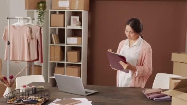 幸せなアジアの女性のオンライン衣料品店のオーナーの肖像画を追跡し 彼女のオフィスに立って クリップボードに書き込み その後 カメラのために笑顔 — ストック動画