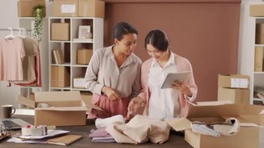 Orta boy Asyalı bir kadın tablete bakarken ve siyahi bir meslektaşla konuşurken çevrimiçi mağazaları için kıyafet paketlerken.