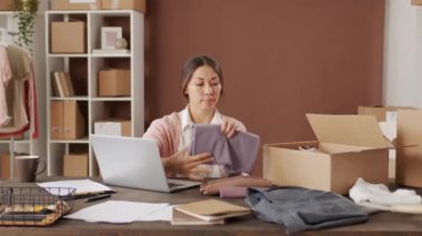 Asyalı bir kadının küçük bir giyim mağazasının ofisindeki masasında otururken ve teslimat siparişini hazırlarken karton kutuya giysi koyarken görüntüsü.