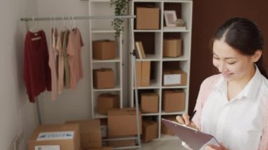 Küçük teslimat şirketinde çalışan ve panoya notlar yazan, sonra da kameraya gülümseyen neşeli Asyalı kadının yüksek açılı portresi.