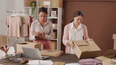 Küçük bir çevrimiçi kıyafet mağazasında çalışan ve teslimat için paketler hazırlayan çok ırklı genç kadınların orta boy fotoğrafları. Kutuları işaretliyorlar ve panoya bir şeyler yazıyorlar.