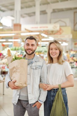 Çiftçilerin pazarında elinde kesekağıdıyla duran gülümseyen genç bir çiftin portresi.