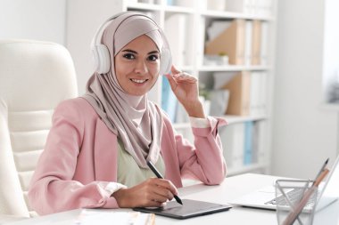 Başörtülü ve kulaklıklı mutlu genç bayan tasarımcı resimlere rötuş yaparken ya da ofiste çizim yaparken sana bakıyor.