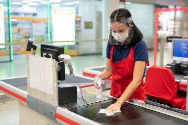 Tanımlanamayan kasiyer kırmızı önlük takıyor. Modern mağazada dezenfektanla kasayı temizliyor.