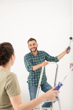 Gülümseyen genç bir kadın erkek arkadaşıyla odayı boyarken duvara boya silen bir kadın.