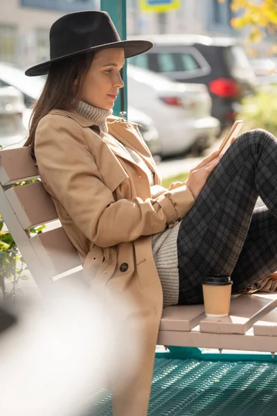 穿着米色风衣 头戴黑色帽子 头戴格子裤子的年轻而文雅 沉稳的黑发女性在智能手机里打滚 在长椅上喝咖啡 — 图库照片