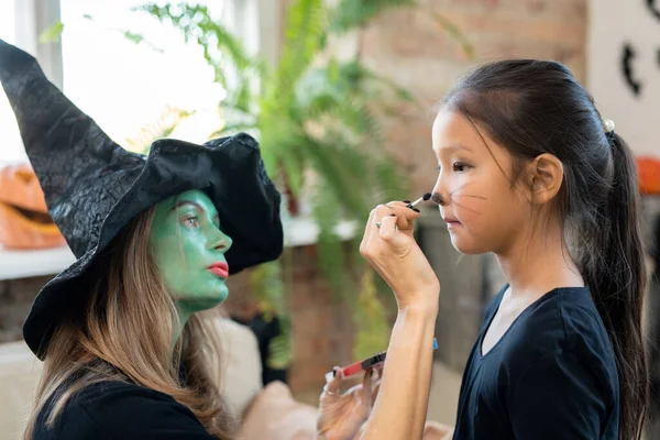 Skoncentrowana Młoda Kobieta Zieloną Twarzą Maluje Wąsy Twarzy Dzieci Przygotowując — Zdjęcie stockowe