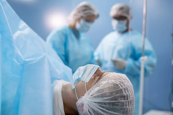 戴防护面具和头戴头饰的年轻男性患者躺在手术台上 与准备医疗器械的医生抗衡 — 图库照片