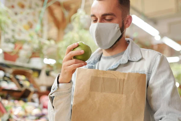 Young male customer in cloth mask standing at food counter and holding paper bag while buying apples at farmers market