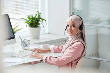 Başarılı genç Müslüman kadın tesettürcü tesettür ve kulaklık tıklayarak fare ve bilgisayar monitörü önünde otururken size bakıyor