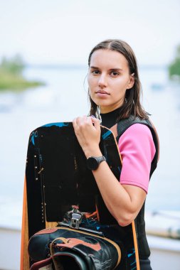 Aktif, uzun saçlı sörf tahtası tutan genç bir bayan sörfçü kameranın önünde doğal ortamda kıyıya karşı duruyor.