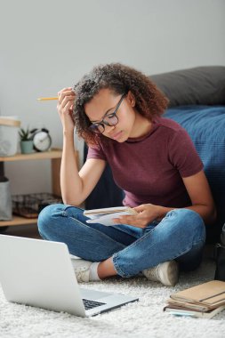 Genç, çapraz bacaklı bir öğrenci dizüstü bilgisayarın önünde yerde oturuyor ve ev ortamında notlarını yeniden yazıyor.