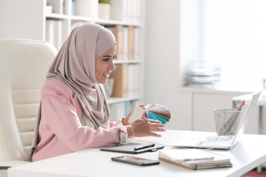 Başörtülü genç Müslüman kadın tasarımcı ve ofiste çalışırken dizüstü bilgisayardaki müşterilerden birine palet gösteren şık günlük kıyafet.