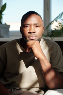 Afrika kökenli genç ve ciddi bir adam kameranın önünde oturmuş ev ortamında bitkilerin olduğu pencereye bakarken sana bakıyor.