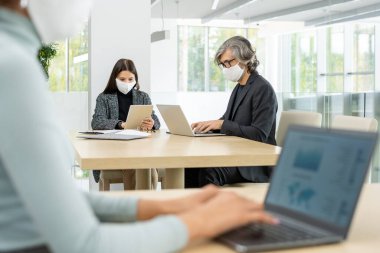 Akıllı günlük kıyafetleri ve koruyucu maskeleri olan iki meşgul iş arkadaşı masa başında oturup geniş ofis ağları kurarken mobil gagdetler kullanıyor.