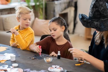 Konsantre tatlı kız masada oturuyor ve öğretmenin kontrolü altında kağıt işçiliğine yapıştırıcı sürüyor. Bu arada sarışın çocuk da sınıftaki zanaatına bakıyor.