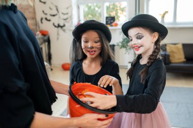 Cadılar Bayramı kostümlü neşeli, çok ırklı kızlar kadınların elinde tuttuğu kovadan şeker topluyorlar. Cadılar Bayramı konseptinde şaka mı şeker mi geleneği.