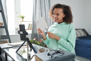 Neşeli genç öğrenci, evdeki akıllı telefon ekranında arkadaşına açıklarken, defterin sayfasındaki notlara işaret ediyor.