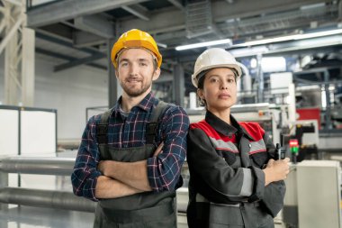 Modern fabrikanın iç veya atölyesine karşı dururken kasklı ve iş elbiseli iki kendine güvenen genç işçi omuz omuza savaşıyor.