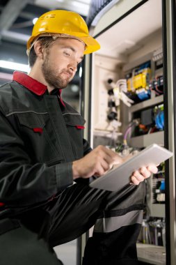 Genç sakallı iş elbisesi mühendisi ve sanayi makinesinin çalışma ayarlarını kontrol ederken tablet içindeki teknik verileri kaydırıyor