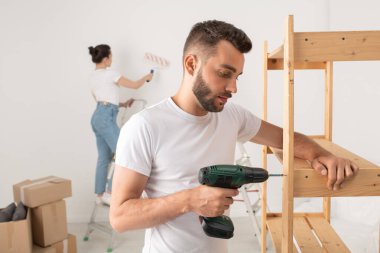 Yeni dairede kablosuz tornavida kullanarak mobilya monte eden konsantre genç sakallı adam.